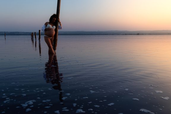 Mujer de pie en agua poco profunda al atardecer, con postes de madera en el fondo.
