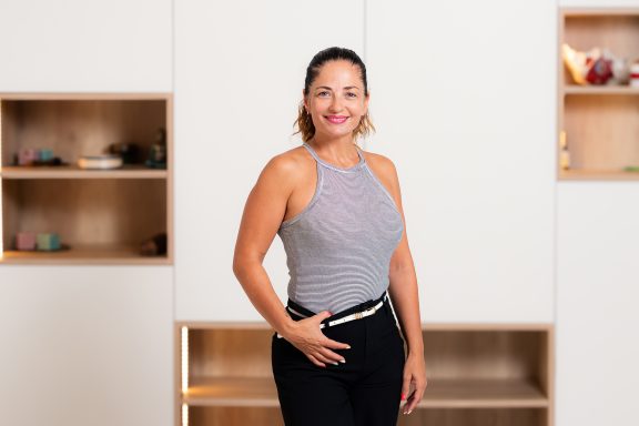 Mujer sonriente de pie en una habitación con estanterías blancas y decorativas.